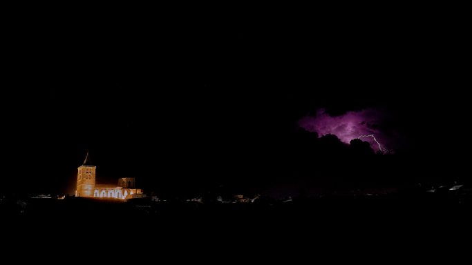 Nit fosca amb una tempesta elèctrica i una església il·luminada al fons.
